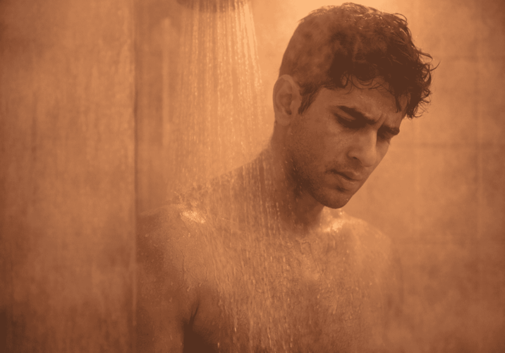 A man standing under a shower head in a steamy, dimly lit bathroom with a warm orange tint.