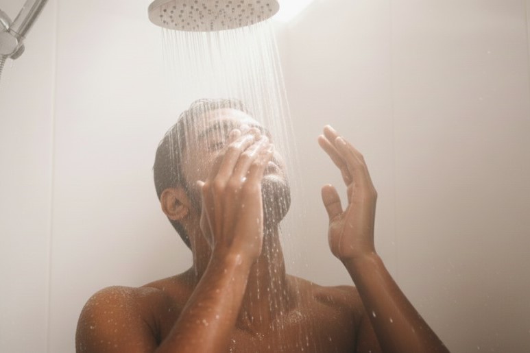 Man washing his face with both hands under a warm shower head; a moment of self-care and gentle cleansing in a steamy bathroom, illustrating perfect water temperature for skincare.