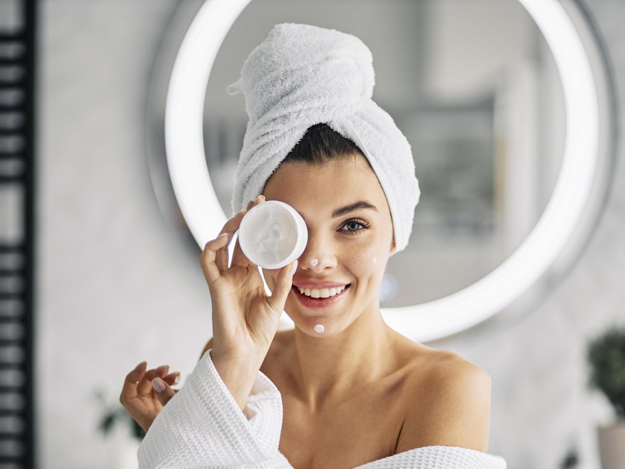A smiling woman in a white bathrobe and head towel playfully holding a jar of face cream over one eye in a bright bathroom.