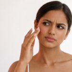 A woman with a concerned expression touching her cheek while looking into a mirror against a plain white background.