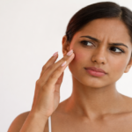 A woman with a concerned expression touching her cheek while looking into a mirror against a plain white background.