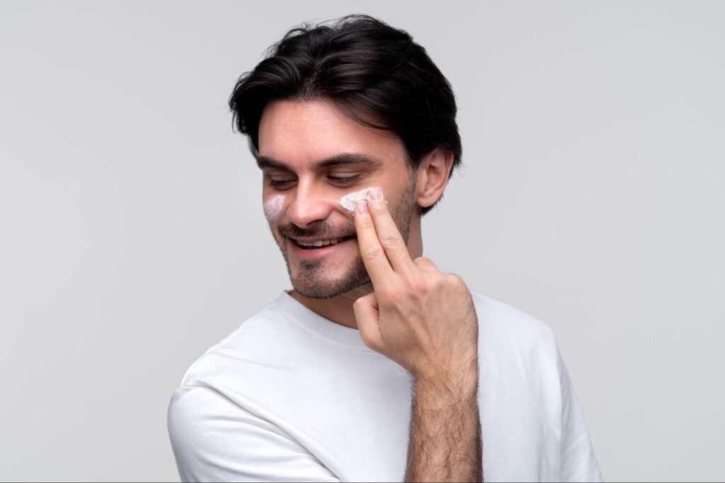 Man smiling while applying face cream to his cheek as part of a daily skincare routine