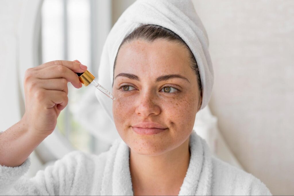 A close-up of a woman with freckled skin, wearing a white bathrobe and a towel wrapped around her hair, applying a clear serum to her cheek using a dropper.