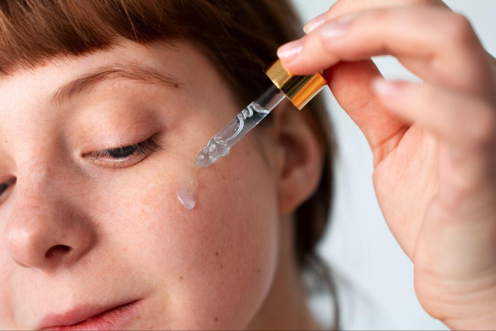 A close-up side profile of a woman with red bangs and freckles applying a droplet of clear serum to her cheek using a glass dropper