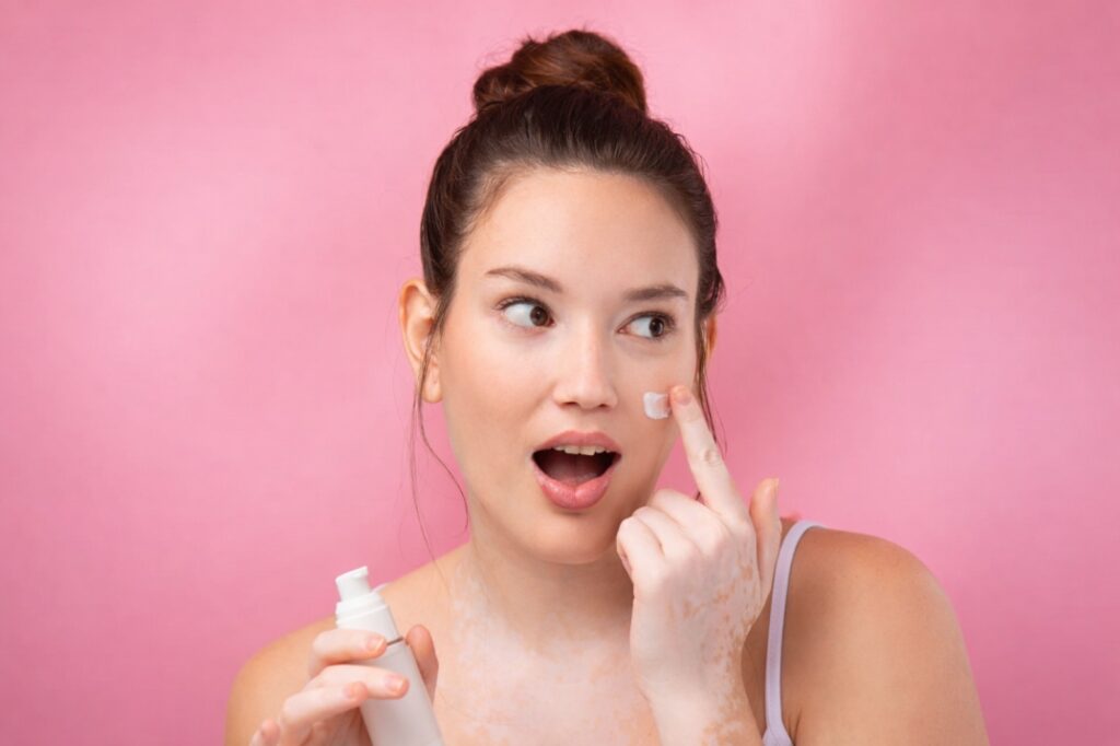A woman with freckled skin, wearing a white bathrobe and a towel wrapped around her hair, holds a glass dropper with a gold cap near her cheek as she looks slightly to the side.