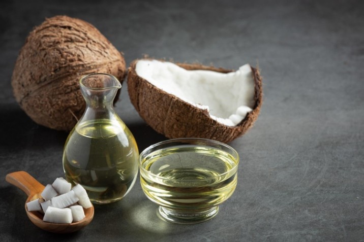 A small glass pitcher and bowl of clear coconut oil alongside fresh coconut halves and pieces on a dark surface.