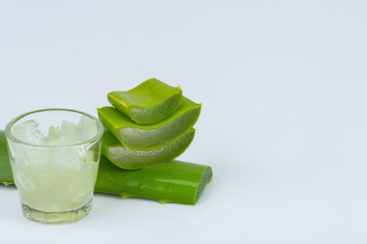Fresh aloe vera leaves stacked next to a glass filled with translucent aloe vera gel against a white background.