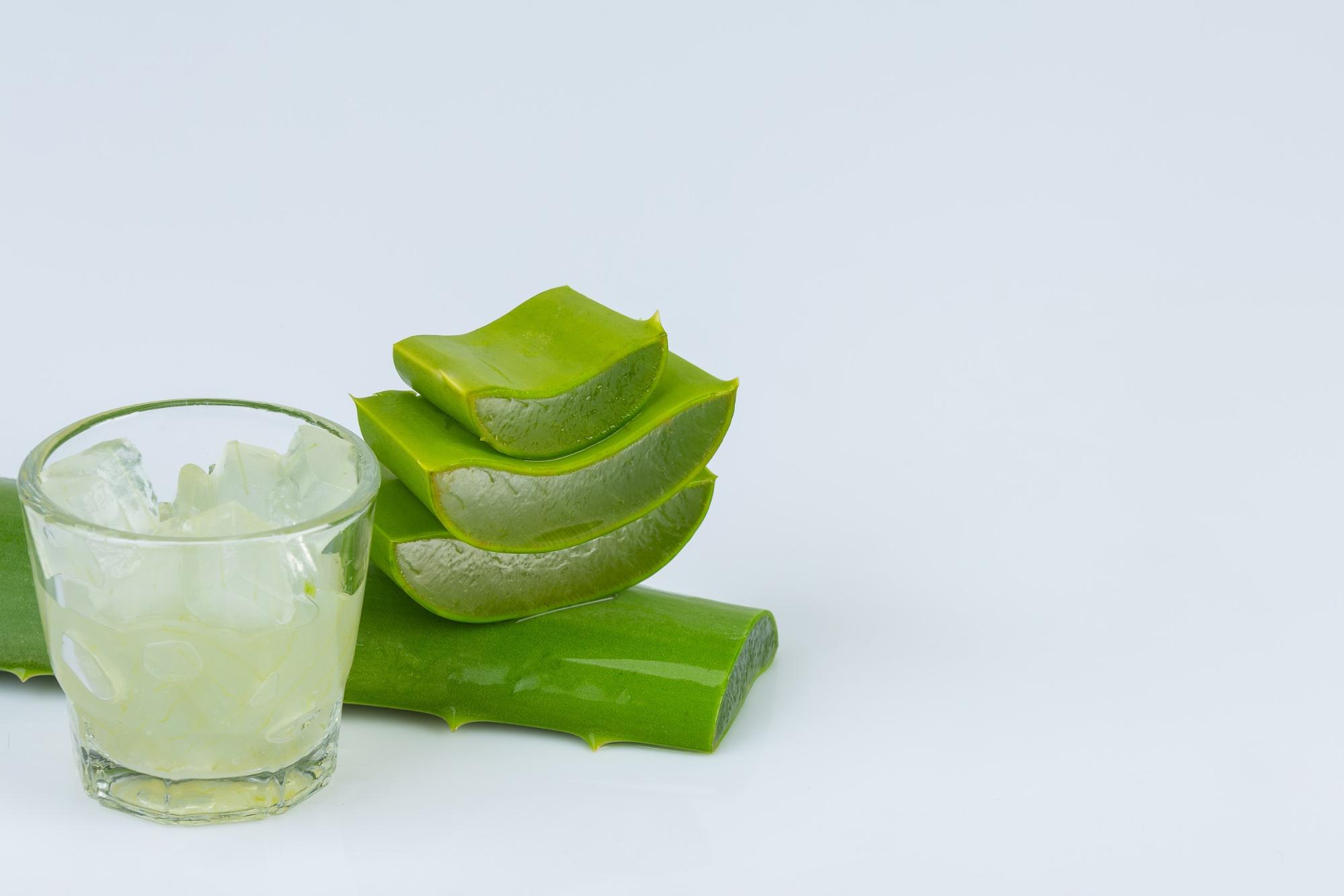 Fresh aloe vera leaves stacked next to a glass filled with translucent aloe vera gel against a white background.