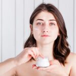 A young woman with brown hair holds a small white jar of face cream. She has a streak of white moisturizer on her cheek and is looking directly at the camera against a white wood-paneled background.
