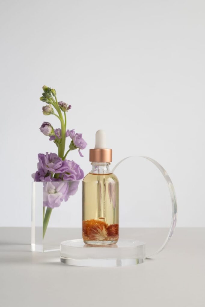 A minimalist product shot of a facial oil bottle with orange botanicals inside, displayed next to purple flowers on a white surface.