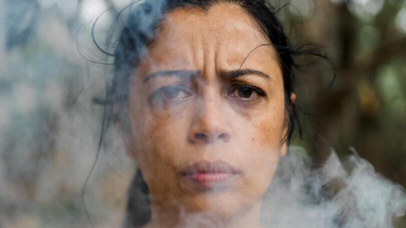 Is-Winter-Pollution-Destroying-Your-Skin? Close-up portrait of a woman with a concerned expression surrounded by thick smoke or smog, representing the impact of air pollution on health.