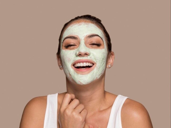 A laughing woman with a light green clay face mask applied to her forehead, nose, and cheeks.