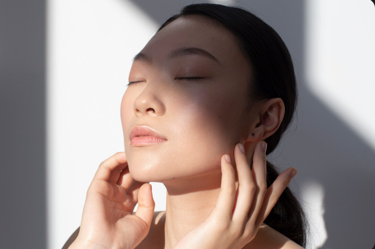 Close-up of a woman with glowing glass skin gently touching her face in natural sunlight to show Korean skincare results.
