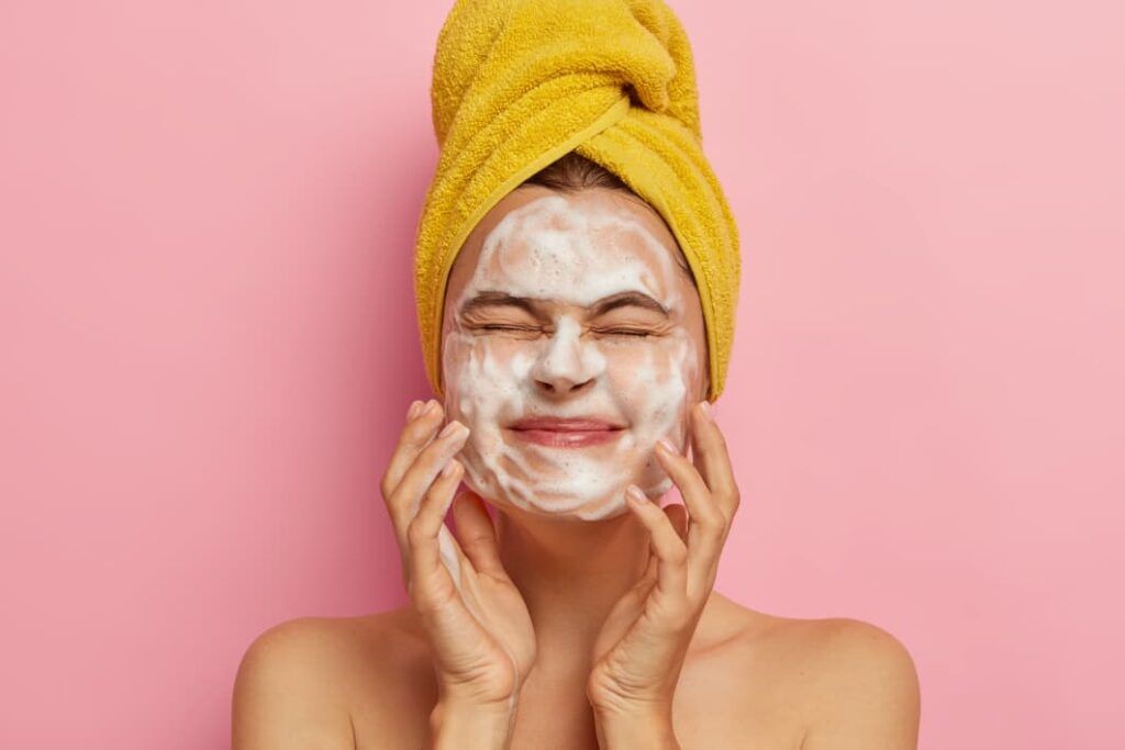 Woman with a yellow head towel applying white foaming cleanser to her face as part of a double cleansing routine.