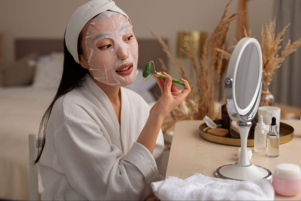 Woman wearing a white sheet mask and using a green jade roller for a facial massage as part of a Korean skincare ritual.