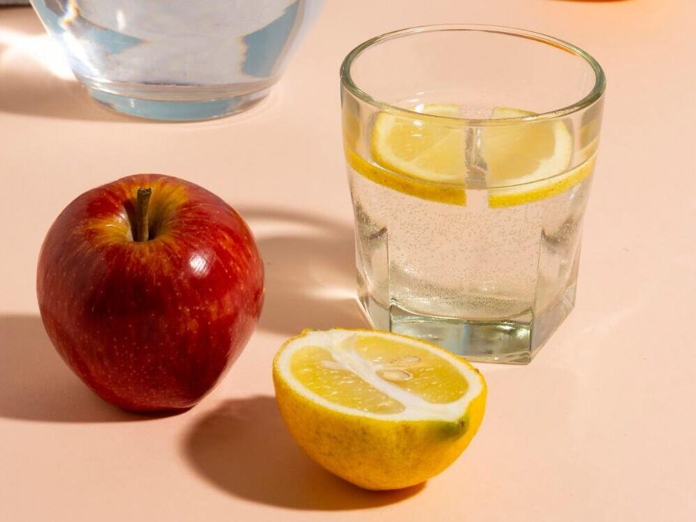 A glass carafe of water next to a glass of sparkling lemon water, a red apple, and a whole orange on a peach and blue background.